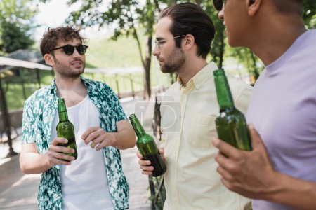 junger, trendiger Mann mit Sonnenbrille, Bier in der Hand und Gespräch mit verschwommenen interrassischen Freunden im Stadtpark
