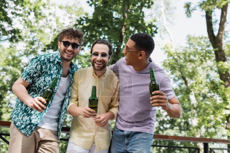 Téléchargez les photos : Joyeux et à la mode interracial amis dans des lunettes de soleil tenant la bière et riant dans le parc vert - en image libre de droit