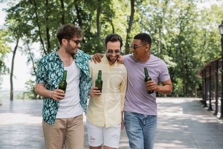 Unbekümmerte und stilvolle interrassische Freunde, die sich beim Sommerspaziergang im Stadtpark umarmen und Bier in der Hand halten