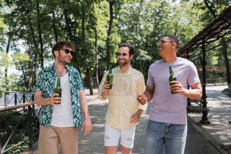 carefree and stylish multiethnic friends holding beer while walking in urban park on summer day