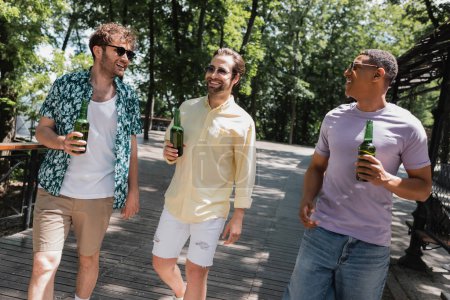Unbekümmerte Multikulti-Freunde im trendigen Sommeroutfit und Sonnenbrille spazieren mit Bier im Stadtpark