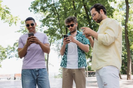 Foto de Hombres multiculturales en traje de verano con estilo y gafas de sol utilizando teléfonos inteligentes en el parque de la ciudad - Imagen libre de derechos