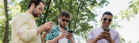 despreocupado hombre afroamericano usando teléfono inteligente cerca de amigos en gafas de sol de moda en el parque verde, pancarta