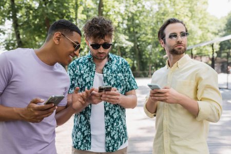 alegre hombre afroamericano señalando con el dedo cerca de amigos de moda utilizando teléfonos celulares en el parque de verano