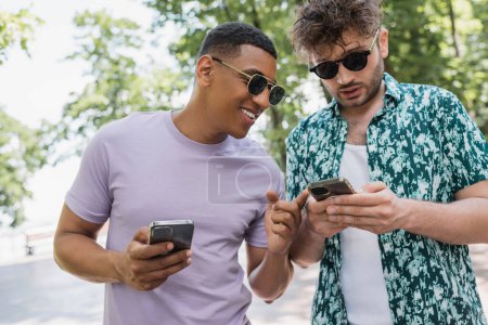 sonriente afroamericano hombre apuntando con el dedo al teléfono inteligente en manos de un amigo de moda en el parque de verano