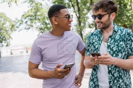 Téléchargez les photos : Homme afro-américain joyeux dans des lunettes de soleil tenant téléphone portable et parler à un ami à la mode dans le parc urbain - en image libre de droit