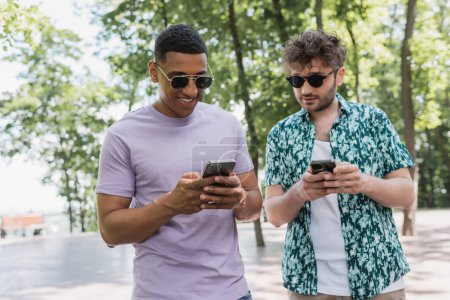 Smiling interracial friends in sunglasses using smartphones in park 
