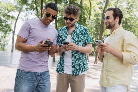 Téléchargez les photos : Un homme afro-américain positif utilisant un smartphone avec des amis proches dans des lunettes de soleil dans le parc - en image libre de droit