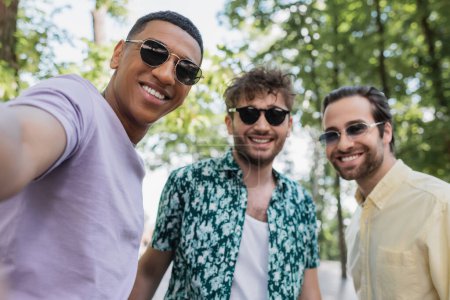 Cheerful african american man in sunglasses standing near blurred friends in summer park 
