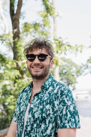 Fröhlicher Mann mit Sonnenbrille und Hemd steht im Sommerpark 