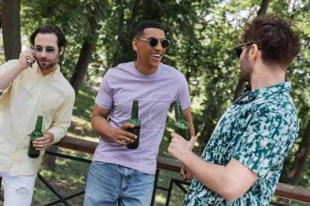 Foto de Hombre afroamericano en gafas de sol sosteniendo botella de cerveza mientras pasa tiempo con amigos en el parque - Imagen libre de derechos