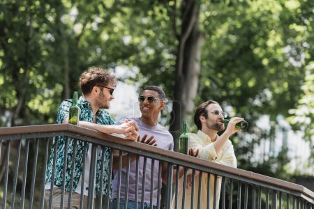 Positive multiethnic friends in sunglasses talking near beer on railing in summer park 