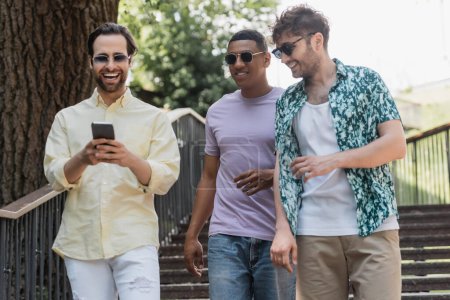 Fröhlicher Mann benutzt Smartphone beim Spazierengehen in der Nähe multiethnischer Freunde auf Treppe im Sommerpark 