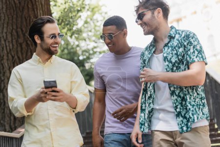 Amigos multiétnicos positivos utilizando el teléfono inteligente mientras camina por las escaleras en el parque en verano 
