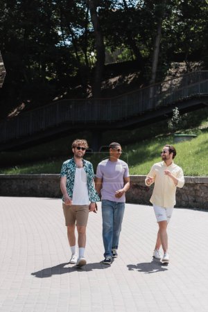 Young man in sunglasses and casual clothes talking to interracial friends while walking in park 