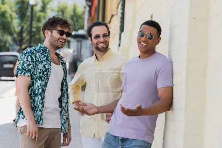 Positive interracial friends in sunglasses standing near building on street in Kyiv 