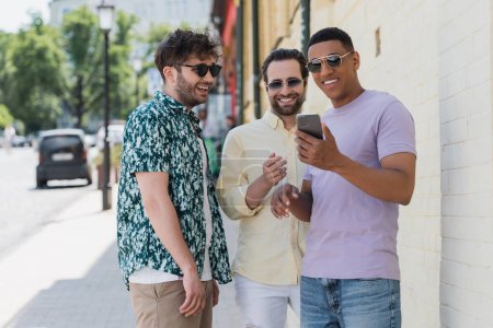 Amis interraciaux insouciants dans les lunettes de soleil en utilisant le téléphone mobile dans la rue à Kiev 