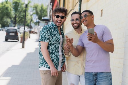 Multiethnische Freunde mit Sonnenbrille schauen weg und halten ihr Smartphone auf der Straße in Kiew 
