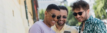 Hommes multiethniques en lunettes de soleil regardant le téléphone portable dans la rue à Kiev en été, bannière 