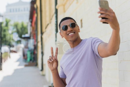 Téléchargez les photos : Souriant homme afro-américain en lunettes de soleil montrant signe de paix tout en prenant selfie dans la rue urbaine floue - en image libre de droit