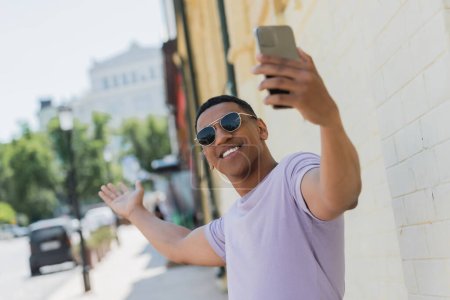 Fröhlicher afrikanisch-amerikanischer Mann mit Sonnenbrille beim Videoanruf auf dem Smartphone auf der Straße in Kiew 
