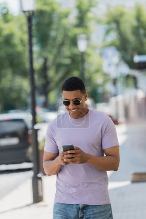 Foto de Hombre afro-americano positivo en gafas de sol usando smartphone en calle urbana en verano - Imagen libre de derechos