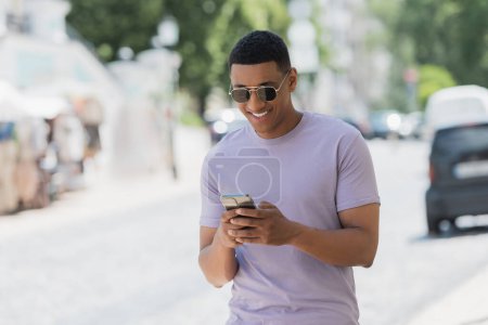 Foto de Hombre afroamericano despreocupado en gafas de sol usando teléfono móvil en la calle urbana - Imagen libre de derechos