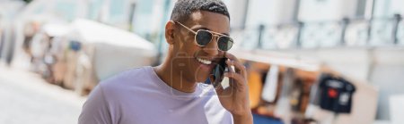 Foto de Hombre afroamericano positivo en gafas de sol hablando en el teléfono inteligente en la calle urbana, bandera - Imagen libre de derechos