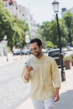 Photo for Cheerful man in shirt and sunglasses using smartphone while walking on urban street - Royalty Free Image