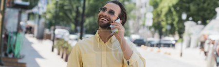 Téléchargez les photos : Homme insouciant en lunettes de soleil parlant sur son téléphone portable dans une rue urbaine floue en été, bannière - en image libre de droit
