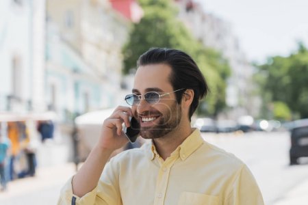 Photo for Cheerful young man in sunglasses talking on smartphone on blurred urban street - Royalty Free Image