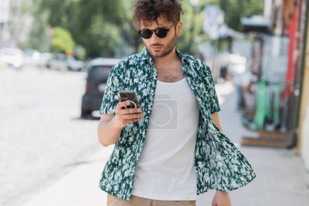 Brünetten Mann mit Sonnenbrille mit Smartphone beim Gehen auf der Stadtstraße 