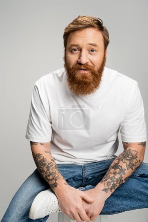 Bearded man in t-shirt and jeans sitting on chair isolated on grey 