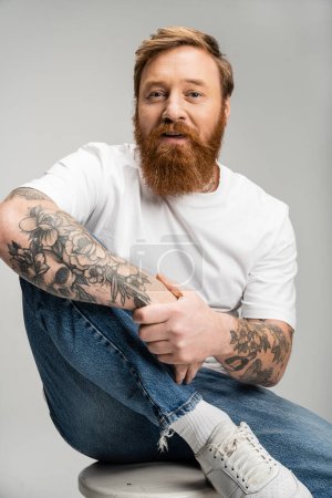 Retrato de hombre tatuado en jeans y camiseta sentado en silla aislado en gris 