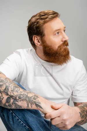 Bearded man in t-shirt and jeans looking away isolated on grey 