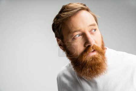 Neugieriger Mann mit Bart trägt weißes T-Shirt und schaut vereinzelt auf grau 