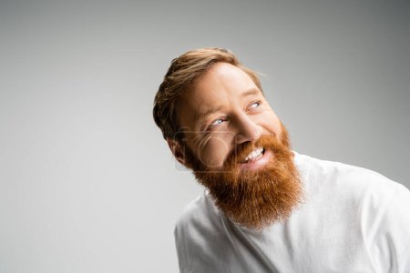 Happy and bearded man in t-shirt looking away isolated on grey 