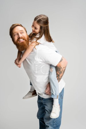 Fröhliches Kind huckepack auf tätowiertem Vater mit Bart isoliert auf grau 