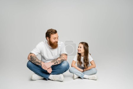 Lächelndes Mädchen im Gespräch mit tätowiertem Vater, während es auf grauem Hintergrund sitzt