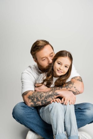 Tätowierter Mann umarmt und küsst Kopf der glücklichen Tochter auf grauem Hintergrund