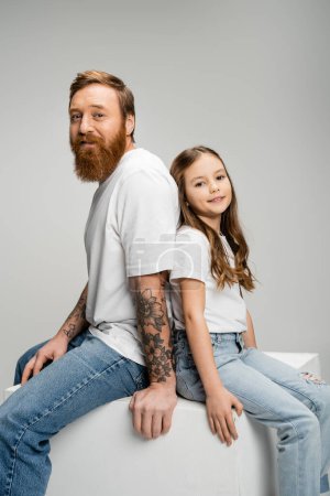Lächelnder Vater und Tochter sitzen Rücken an Rücken auf Würfel isoliert auf grau  