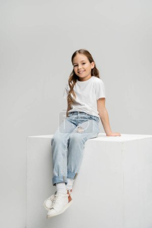 Téléchargez les photos : Enfant joyeux en t-shirt et jeans assis sur cube isolé sur gris - en image libre de droit
