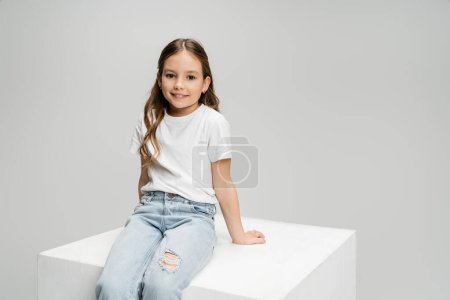 Smiling preteen girl in t-shirt and jeans sitting on cube isolated on grey  