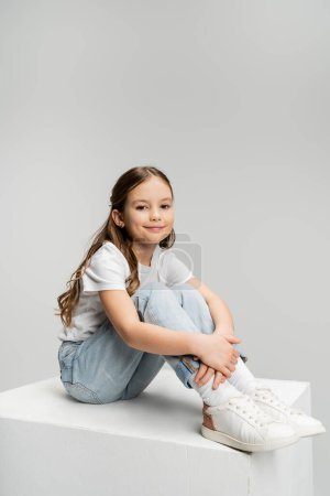 Smiling preteen child in casual clothes sitting on white cube isolated on grey  
