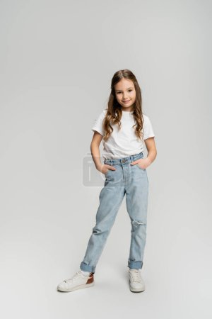 Téléchargez les photos : Pleine longueur de l'enfant positif en jeans et t-shirt posant sur fond gris - en image libre de droit