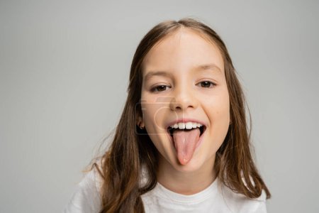 Niño preadolescente sobresaliendo lengua en la cámara aislado en gris  