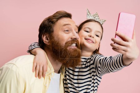 Foto de Chica sonriente con diadema de corona abrazando a papá barbudo mientras toma selfie aislado en rosa - Imagen libre de derechos