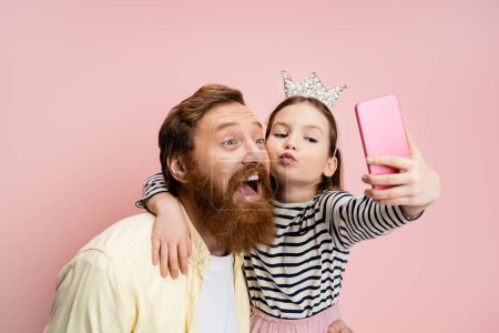 Foto de Chica preadolescente con diadema de corona haciendo pucheros labios y tomando selfie con papá barbudo aislado en rosa - Imagen libre de derechos