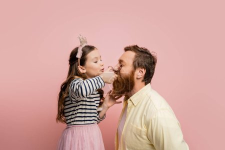 Preteen girl with crown headband curling eyelashes of father isolated on pink  