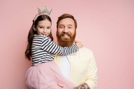 Positive und tätowierte Vater hält Tochter mit Krone Stirnband isoliert auf rosa  